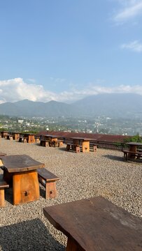 Outdoor cafe seating with wooden tables and benches on a gravel patio, overlooking a scenic valley and mountain range under a partly cloudy sky.