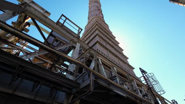 Industrial processing plant in backlight with massive chimneys, storage tanks, pipelines and complex machinery, showcasing large-scale production infrastructure under a bright blue sky.