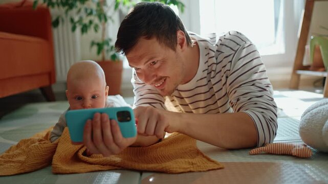 Young dad and his curious newborn son lying on the floor watching cartoons on a smartphone, highlighting modern parenting and the early introduction of digital devices to children