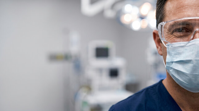 Close-up half face portrait of a male surgeon wearing protective safety goggles and a surgical mask inside a modern hospital operating theater with copy space on the left.