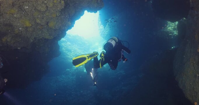 Scuba divers exploring a dramatic underwater rocky canyon, surrounded by clear blue water, marine life and sunlight filtering through the submerged rock formations.