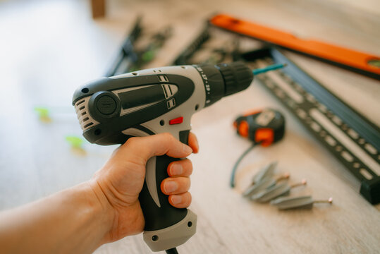 Person holding a power drill, preparing for a home improvement project, with a tv mounting bracket, tape measure, and wall anchors visible on the floor, ready for installation work