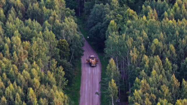 Aerial view of a combine harvester driving along a narrow dirt road through a dense forest in Latvija.