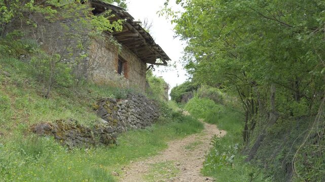 A quaint dirt path meanders through lush green hills, passing an ancient stone house in Lin, Albania. Tilt Up Shot