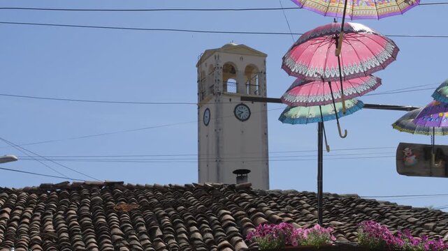 Capturing the historic Elbasan Clock Tower in Albania, a sunny tilt-up shot reveals decorative umbrellas and a clear blue sky.