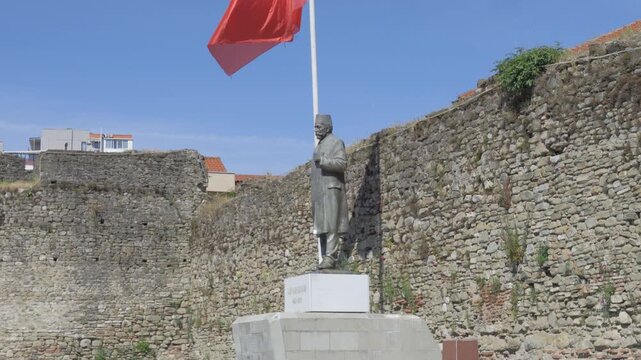 Lef Nosi statue beneath a waving Albanian flag beside old stone fortress walls in Elbasan.