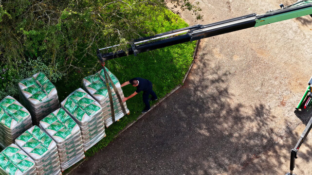 Aerial view of a worker operating a crane to lift a pallet of bagged materials on a paved surface near a grassy area in Latvija.