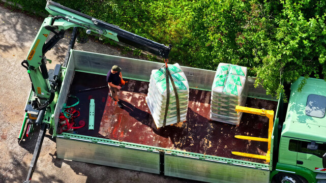 Aerial view of a worker loading pallets of bagged construction materials onto a flatbed truck with a crane in Latvija.