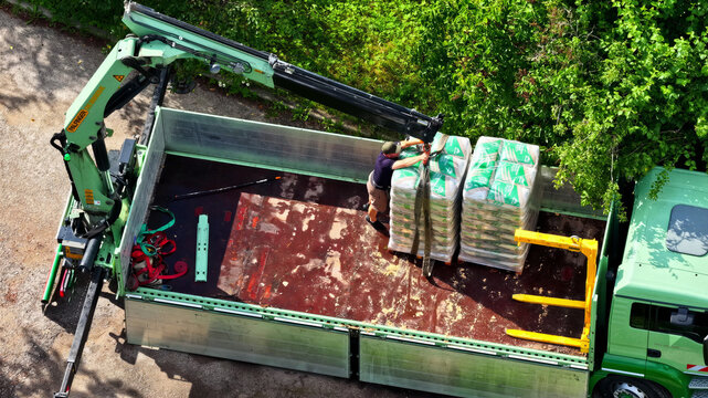 Aerial view of a worker loading pallets of bagged material onto a green flatbed truck with a crane in Latvija.