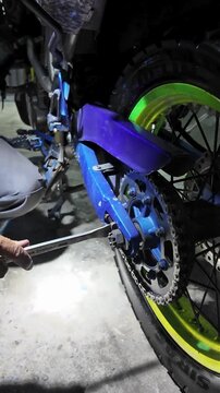 close-up mechanic using spanner wrench to adjust drive chain tension on dirt bike. routine motorcycle maintenance or repair in a garage workshop setting.at night