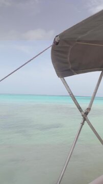 Vertical smartphone video panning right from inside a white motorboat under a bimini top canopy canopy shade, showcasing the calm, multi-toned turquoise waters of a remote Caribbean beach
