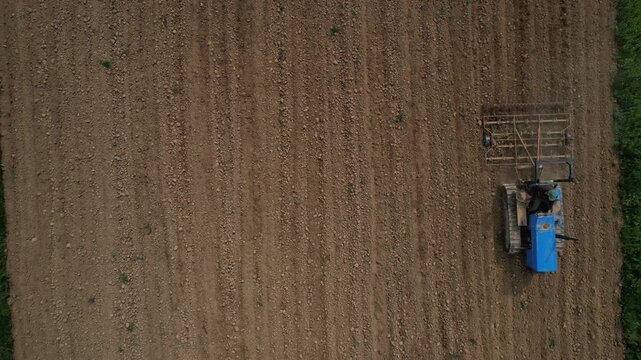 Tractor pulls a wide tillage implement across a freshly worked agricultural field, leaving parallel soil rows and a uniform cultivated surface, top down tracking flight