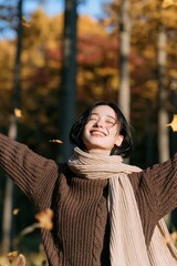 Fototapeta premium Smiling woman in autumn forest background with cozy sweater and scarf for fall banner and seasonal lifestyle design