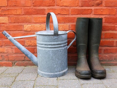 Zinc watering can and garden boots in front of a red brick wall.