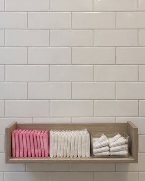 Period poverty concept shows Wooden shelf mounted on a white tile wall holding pink and white sanitary pads and folded cloths.