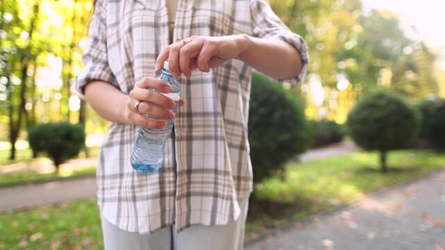 Female hands open plastic water bottle against soft park bokeh. Closeup captures hydration moment and concern about microplastic exposure.