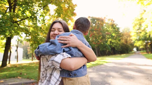 Mother tightly hugs son in autumn park while child appears reluctant to cuddle. Loving embrace continues despite his unhappy mood.