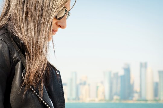 Close up profile young woman brown hair, sunglasses, black leather jacket. Blurred Bay skyline, waterfront corniche background. Soft sunlight, outdoor portrait