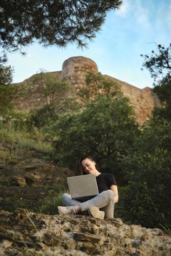 Digital nomad works remotely on a laptop, set against a scenic backdrop of lush greenery and historic architecture in a rocky area