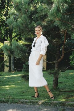 A model stands confidently in a striking white shirt and skirt with fringed details, surrounded by vibrant greenery