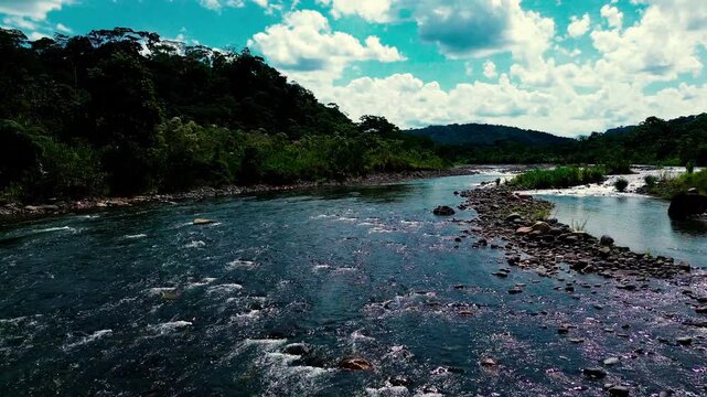 A drone view looks down on a narrow footbridge spanning a rocky river with whitewater, gravel bars and pale exposed banks. Dirt paths lead to the crossing through dense green tropical forest in Ecuado