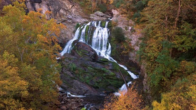 Powerful cascade waterfall flows through rocky gorge surrounded by lush forest and vibrant autumn trees. Mountain river creates tranquil natural scenery.