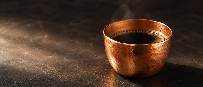 Fresh hot black coffee served in a rustic hammered copper cup with gentle steam on a dark textured marble surface.
