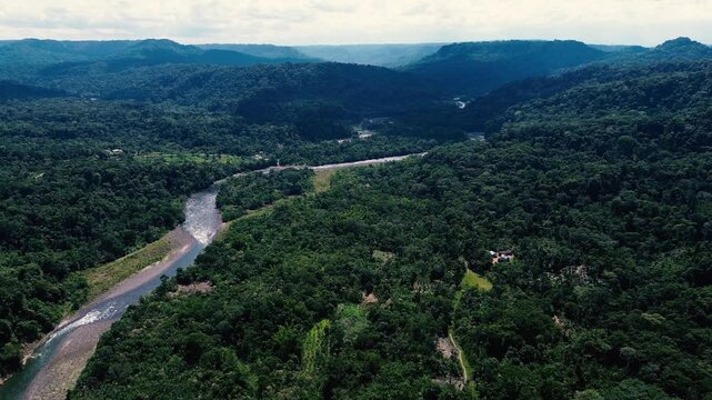A drone aerial view of a shallow river flowing around broad pebble and stone bars, with small rapids and dense tropical forest along the banks. Wooded hills and a bright, cloud-filled sky frame the ri