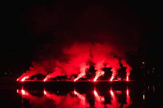 City festival nightscape featuring dense red firework smoke and bright pyrotechnic illumination reflecting across a dark urban body of water.