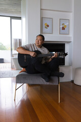 Fototapeta premium Senior Asian man sitting on bench playing black acoustic guitar in modern living room