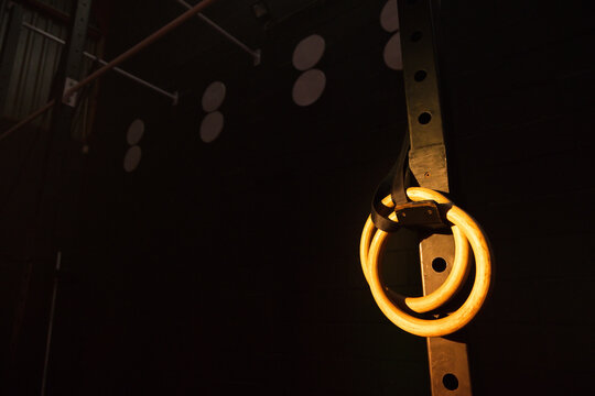Wooden gymnastic rings are hanging from black straps on metal upright in dim gym, casting shadows