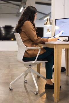 Woman wearing brown blazer sitting at desk in office holding pages and pen viewing monitor charts