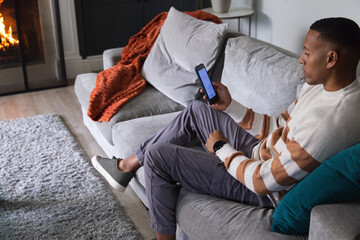 Fototapeta premium African American man sitting on gray sofa in living room holding phone, orange blanket, copy space