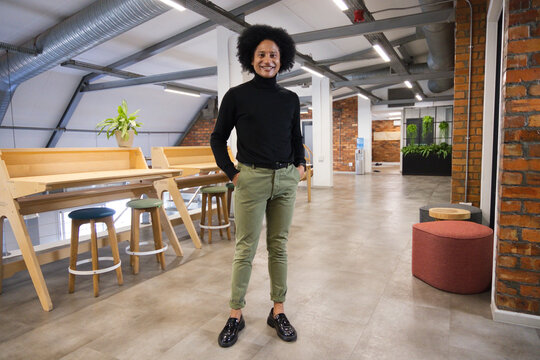 Adult African man standing in loft office wearing black turtleneck and olive chinos near benches