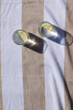 Two clear glasses are resting on wide striped towel, with ice, lemon wedges and sunlit shadows