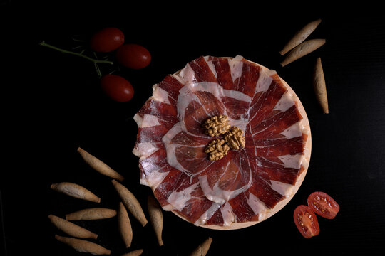 Top view of sliced iberian ham on a wooden board decorated with walnuts, breadsticks, and tomatoes over a black background, gourmet spanish appetizer concept