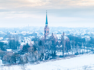 Zimowa panorama Nowej Wsi Królewskiej z lotu ptaka. Ceglany kościół z wysoką, zieloną wieżą otoczony ośnieżonymi drzewami i zabudową miejską.  © Henryk Niestrój