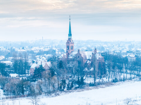 Zimowa panorama Nowej Wsi Kr&oacute;lewskiej z lotu ptaka. Ceglany kości&oacute;ł z wysoką, zieloną wieżą otoczony ośnieżonymi drzewami i zabudową miejską. 