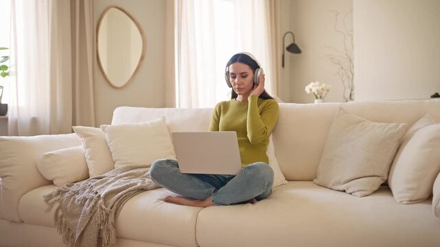 Young Caucasian pretty woman wearing headphones working on laptop while sitting cross legged on sofa. Female remote worker concentrating on computer in cozy bright modern living room.