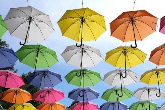 Rows of open umbrellas in white, yellow, orange, green, blue and pink suspended overhead against a clear sky. Low-angle view of dozens of brightly colored umbrellas hung in neat rows abo