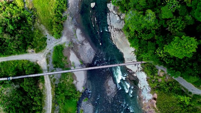 An overhead drone view shows a narrow bridge spanning a rocky river bordered by dense green forest in Ecuador's Oriente. Gravel banks, branching dirt paths and small rapids shape the tropical river la