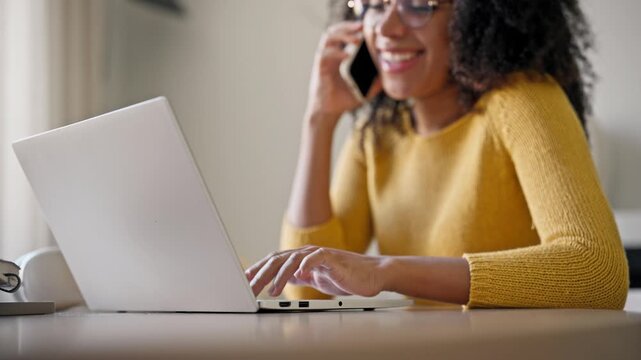 Young African American woman talking on smartphone and typing on laptop at table. Smiling female multitasking while working remotely and having phone conversation from comfortable home.