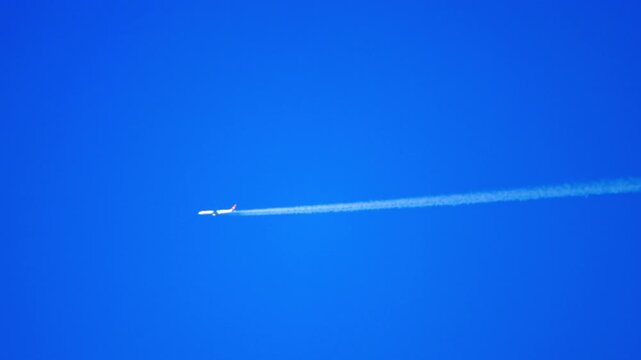 Commercial airliner cruises high in clear blue sky leaving long white contrail, telephoto tracking shot