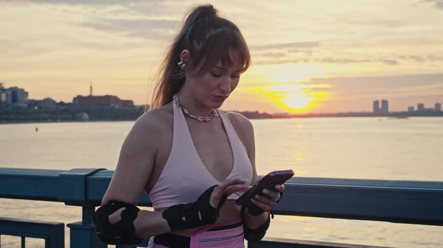 Young girl scrolls navigation application on mobile. Beautiful rollerblader explores local area looking around promenade during golden hour over wide picturesque river surface.