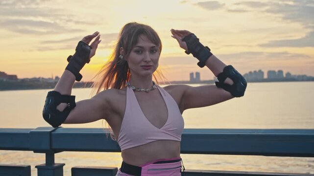 Beautiful girl fixes hair looking at camera at sunset. She stands on river promenade wearing protective gear enjoying warm summer lifestyle vibe after active rollerblading outdoor session.
