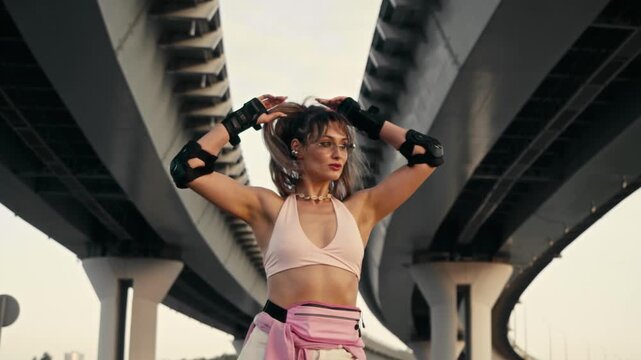 Young woman arrives on rollerblades fixing hair under bridge. Sporty female skater enjoys active urban lifestyle during warm summer day wearing protective gear.