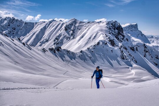 Skier touring across sunlit alpine snowfield with backpack and poles leaving curved tracks under jagged mountain ridges beneath clear blue sky in winter daylight. Davos Klosters Switzerland.