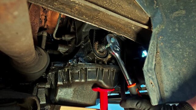 A car mechanic in gloves uses a socket ratchet wrench to tighten a crankshaft pulley bolt during engine repair under an automobile, close-up.