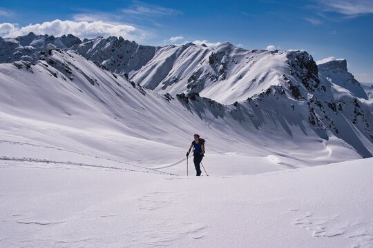 Skier touring across sunlit alpine snowfield with backpack and poles leaving curved tracks under jagged mountain ridges beneath clear blue sky in winter daylight. Davos Klosters Switzerland.