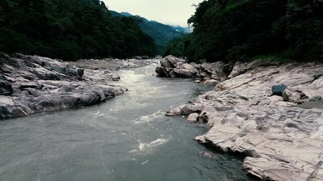 An aerial view looks down on a river flowing around pale rock outcrops and gravel bars, with dense green tropical forest enclosing the channel. Filmed in Ecuador's Oriente region of South America, the